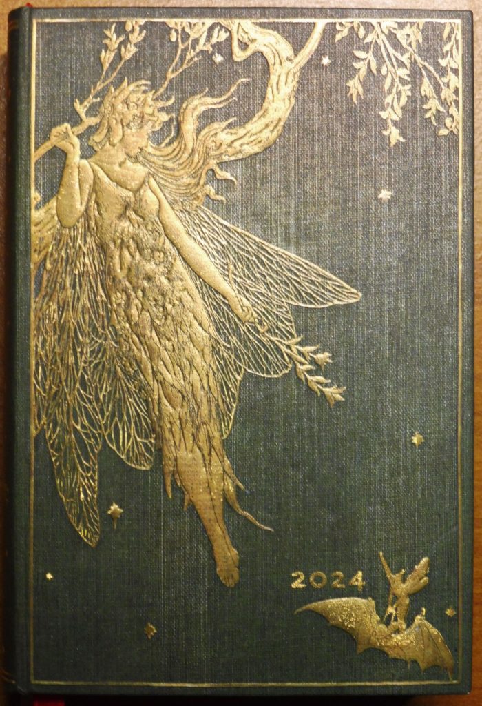 In Leinenoptik gebundener Kalender, dunkelgr&uuml;n, mit erhaben gepr&auml;gtem goldenen Dekor im Jugendstil: Eine Fee wie aus griechischer Mythologie mit Fl&uuml;geln und Zweigen in den H&auml;nden; sie schaut nach rechts, aus ihren Haaren entspringt nach oben ein Baumstamm mit Zweigen und Bl&auml;ttern. Unten rechts in der Ecke eine Fledermaus und direkt vor/&uuml;ber ihr eine nicht erkennbare Figur, vielleicht eine Elfe, sowie die Zahl 2024. Es sind &uuml;ber das gesamte Bild einige Sterne verstreut.