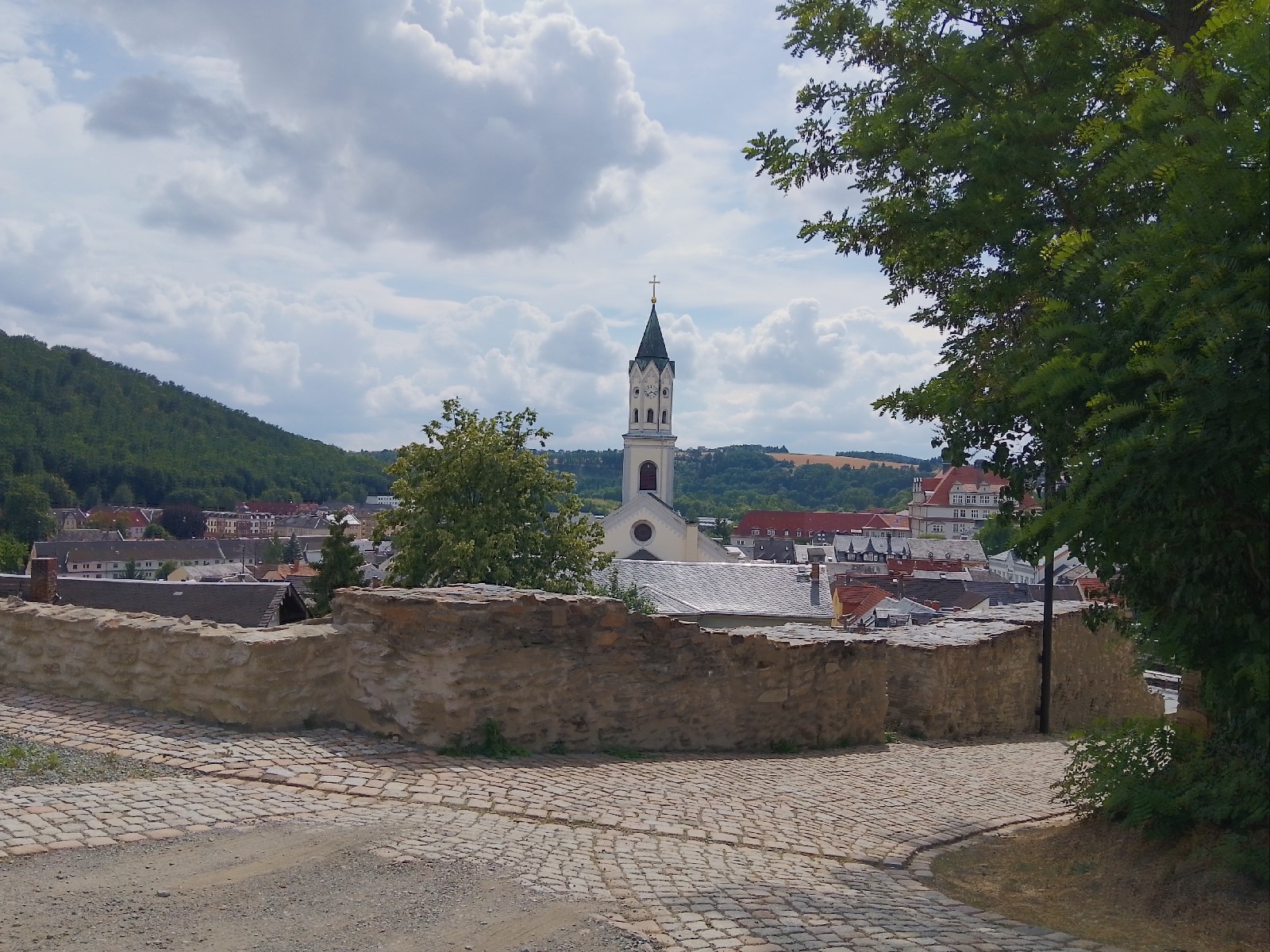 Ausblick. Zentral im Bild der helle Kirhturm mit Uhr hinter einer niedrigen Burgmauer. Rechter Bildrand wird von enem Laubbaum dominiert.