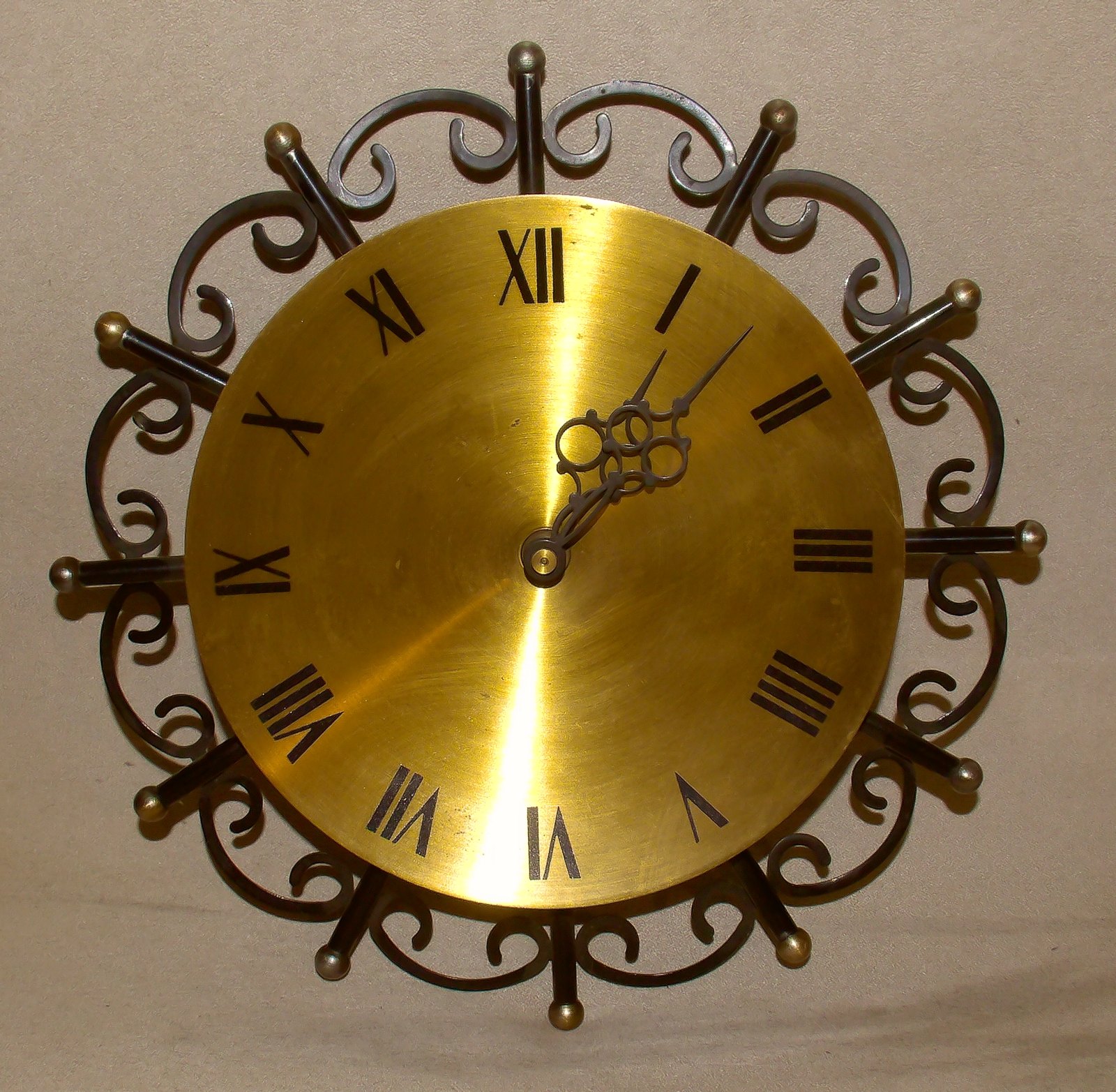 Runde Wanduhr. Vor zwölfstrahligem ornamentalem, dunklem Metall ein Messingzifferblatt mit Römischen Ziffern, die Zeiger mit Verschnörkelung. Runde Wanduhr. Vor zwölfstrahligem ornamentalem, dunklem Metall ein Messingzifferblatt mit Römischen Ziffern, die Zeiger mit Verschnörkelung.